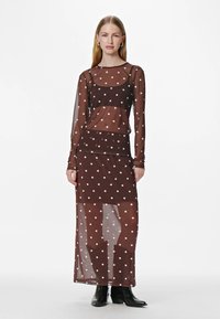 Vestito in rete marrone con pois bianchi, maniche lunghe, gonna alla caviglia, tessuto trasparente, design a strati e stivaletti neri alla caviglia.