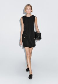 Schwarzes ärmelloses Kleid mit Seitentaschen und goldenen Akzenten, kombiniert mit einer kleinen schwarzen Handtasche und schwarzen Slippern. Glatte Stoffstruktur.