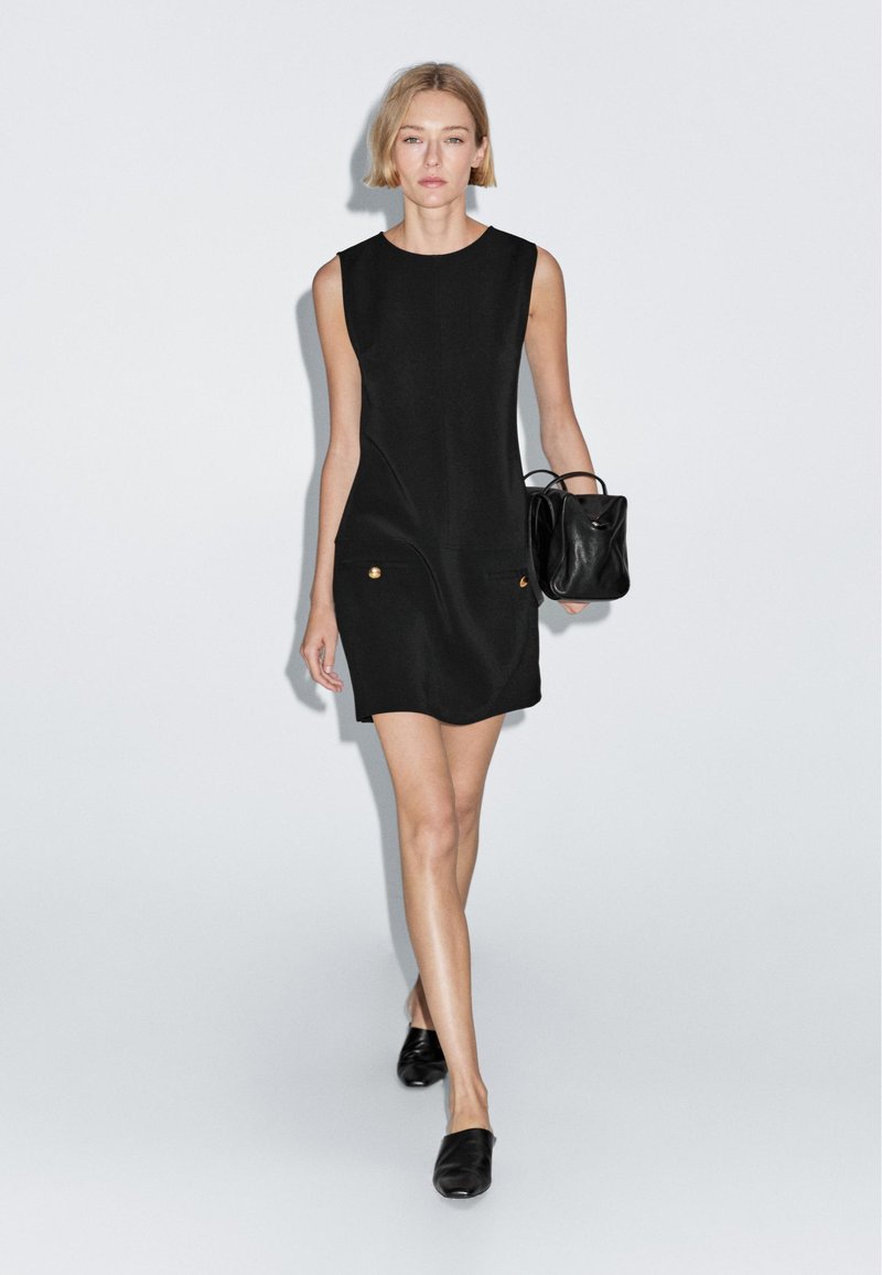 Schwarzes ärmelloses Kleid mit Seitentaschen und goldenen Akzenten, kombiniert mit einer kleinen schwarzen Handtasche und schwarzen Slippern. Glatte Stoffstruktur.