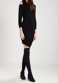Czarna sukienka typu off-shoulder z długimi rękawami i dopasowanym krojem, z marszczeniem na dole. Połączona z czarnymi botkami sięgającymi za kolano.