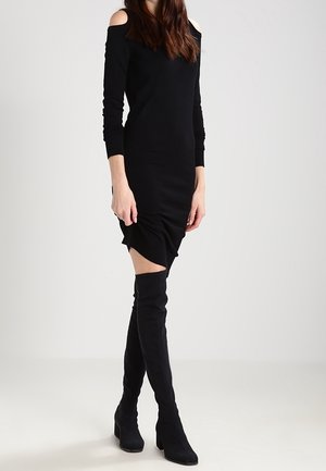 Abito nero off-shoulder con maniche lunghe e silhouette aderente, caratterizzato da ruches all'orlo. Abbinato a stivali neri sopra il ginocchio.