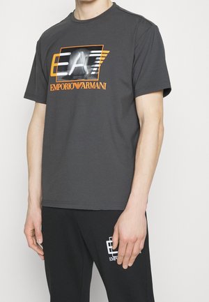 T-shirt en coton gris avec un graphisme E.A. en noir et orange. Manches courtes et col rond, associé à un pantalon noir avec un détail logo.