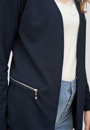 Cardigan blu navy con trama, apertura frontale, maniche lunghe e tasca laterale con zip in metallo argentato. Abbinato a una maglietta bianca.