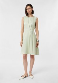 Ärmelloses, hellgrünes Kleid mit gerafftem Taillenbereich, knielanger Saum und glatter Textur. Kombiniert mit weißen, spitzen Ballerinas und einem silbernen Armband.