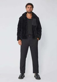 Un homme se tenant debout, portant une veste noire en peluche, une chemise noire, un pantalon noir et des chaussures en cuir noires devant un fond clair.