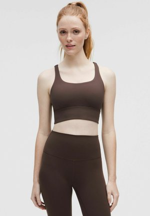 Młoda kobieta z czerwonymi włosami związanymi w kucyk, ubrana w brązowy krótki top i legginsy z wysokim stanem na jednolitym tle.