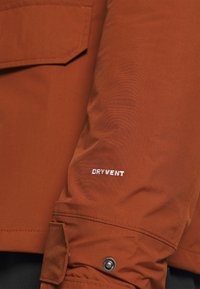Voděodolná bunda v rezavé barvě; má hladkou texturu, kapsu na rukávu a viditelné bílé logo "DRYVENT".
