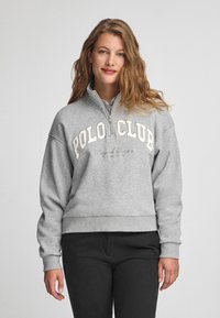 Grå zip-up sweatshirt med hög krage, med "POLO CLUB" i vit text över bröstet och ribbad nederkant. Bärs med svarta byxor.