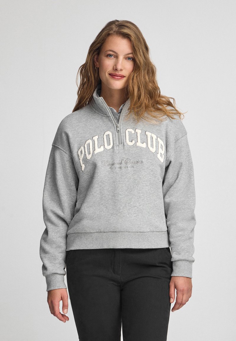 Grå zip-up sweatshirt med hög krage, med "POLO CLUB" i vit text över bröstet och ribbad nederkant. Bärs med svarta byxor.