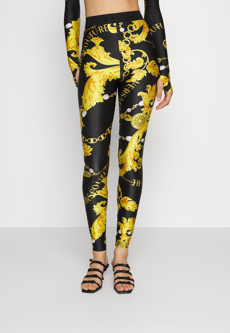 Žena nosi crne tajice s živopisnim žutim baroknim i lančanim uzorkom, u paru s crnim sandalama s remenčićima i odgovarajućom majicom s dugim rukavima.