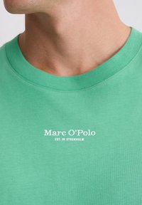 Zielona bawełniana koszulka z okrągłym dekoltem, z białym nadrukiem logo i napisem "Marc O'Polo EST. IN STOCKHOLM" na przodzie. Gładka faktura.