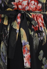 Blommig plisserad klänning med svart bas, prydd med livfulla röda, gula och blå blommor, accentuerad av ett matchande knytbälte vid midjan.