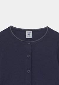 Mörkblå cardigan med ett texturerat mönster, rund halsringning och tre knappar. Har en kontrasterande mörk söm längs kragen.