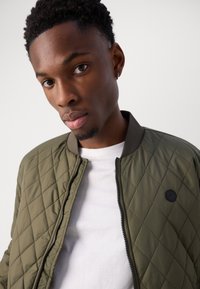 Veste bomber matelassée de couleur olive avec une fermeture éclair et un col sombre, portée sur un T-shirt blanc. Présente un détail de logo noir rond.
