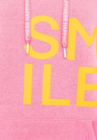 Rosa Sweatshirt mit fettem gelben Schriftzug. Kordeln mit dem Text "GLÜCKSELE". Weicher Stoff, lässiges Design und Kängurutasche.