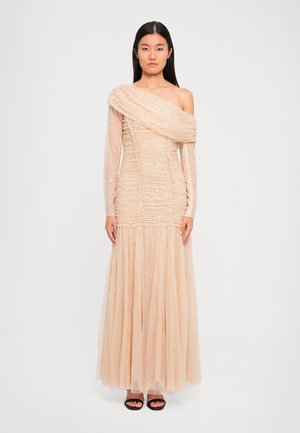 Vrouw in een lange, beige, gerimpelde tule jurk met één off-the-shoulder mouw en subtiele pailletjes, gecombineerd met zwarte open teen hakken.
