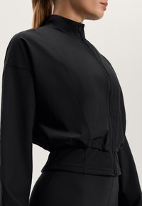 Černá zipová sportovní bunda vyrobená z hladké tkaniny. Má vysoký límec, dlouhé rukávy a přiléhavý pas s nařaseným efektem.