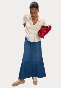 Polo en tricot beige avec un col, associé à une longue jupe en denim et des baskets tan ; tenant un sac en osier rouge.