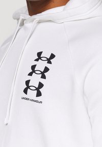 Felpa bianca con tre loghi neri verticali di Under Armour e il nome del marchio sul petto, indossata da una persona con pelle marrone.