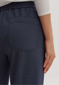 Mörkblå texturerade byxor med en bakficka och resår i midjan, med en delvis synlig hand som vilar på sidan.