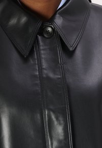 Cappotto in pelle nera con ampio colletto, chiusura centrale a bottoni, superficie liscia e cuciture visibili lungo la patta.
