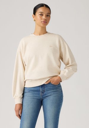 Frau steht mit der Hand in der Tasche, trägt einen beigen Pullover und blaue Jeans vor einem schlichten hellen Hintergrund.