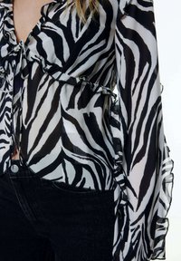 Bluse mit Zebra-Muster in Schwarz und Weiß, aus leichtem, transparentem Stoff mit gerafften Kanten und langen Ärmeln. Kombiniert mit schwarzen Jeans.