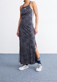 Leopardikuvioinen maksimekko harmaasta kankaasta, jossa on sivuhalkiot ja valkoiset pystysuorat raidat. Yhdistetty harmaisiin lenkkareihin ja siniraidallisiin sukkiin.