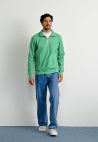 Homem a caminhar, vestindo um suéter verde claro de meia-zíper, jeans azuis e ténis brancos, contra um fundo branco simples.