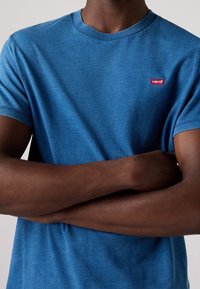 Modrá bavlněná tričko s kruhovým výstřihem, krátkými rukávy a malým červeným logem Levi's na hrudi. Hladká textura, volný střih.