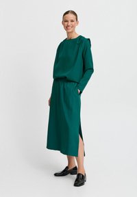 Robe verte à manches longues avec une coupe décontractée, taille élastique, fentes latérales et texture douce ; associée à des mocassins noirs.
