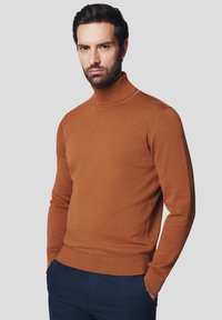 Brązowy sweter typu golf w miękkiej dzianinie, dopasowany krój z prążkowanym dołem i mankietami, noszony na ciemnoniebieskich spodniach.