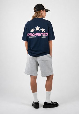 Person trägt eine schwarz-weiße Kappe, ein dunkelblaues T-Shirt mit dem Aufdruck "PROHIBITED" und Sternen in Pink und Weiß, graue Shorts, weiße Socken und schwarze Schuhe.