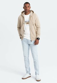 Sudadera cremosa con cremallera, camiseta gráfica blanca, jeans de un azul claro y zapatillas blancas con suelas marrones, de pie sobre un fondo neutro.