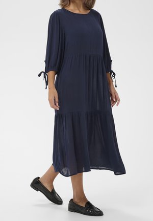 Donna che indossa un abito midi blu navy con maniche a sbuffo annodate ai gomiti e mocassini in vernice nera, in piedi su sfondo bianco.