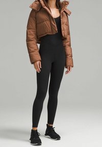 Chaqueta de plumas corta marrón con capucha, combinada con un body negro y leggins negros. Tejido texturizado y zapatillas deportivas negras.