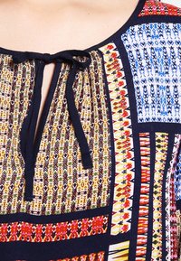 Blusa multicolore con audaci motivi geometrici. Presenta un collo a chiave con fiocco. Realizzata in tessuto leggero, principalmente blu navy e tonalità brillanti.
