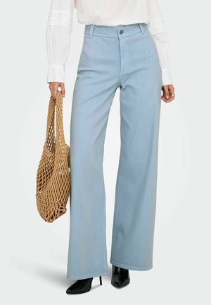 Žena v svetlomodrých nohaviciach s širokými nohavicami, bielej blúzke s dlhými rukávmi a krajkovými detailmi, čiernych špicatých lodičkách, drží tkanú hnedú kabelku.