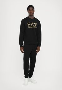 Svart hettegenser med gull "EA7 Emporio Armani" logo, kombinert med svarte joggebukser og hvite sneakers. Glatt tekstur, avslappet design.