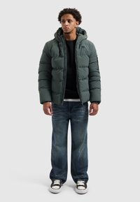 Jonge man in een donkergroene pufferjack met capuchon, zwarte sweatshirt, losse blauwe jeans en zwart-witte sneakers tegen een effen achtergrond.