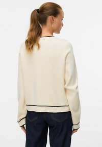 Krēmkrāsas džemperis ar apaļu izgriezumu, garām piedurknēm un melnu apmali ap apakšu un piedurknēm, izgatavots no mīksta materiāla, kopā ar tumšām džinsa biksēm.