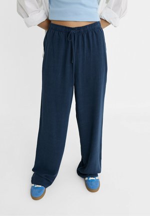 Personne portant un pantalon ample bleu marine à cordon, des baskets bleues avec des détails blancs, et un top court bleu clair avec une chemise blanche par-dessus.
