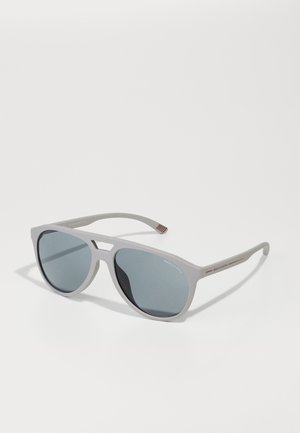 Ochelari de soare din plastic gri, cu un design geometric, lentile întunecate și accente roșii pe brate, cu un finisaj mat.
