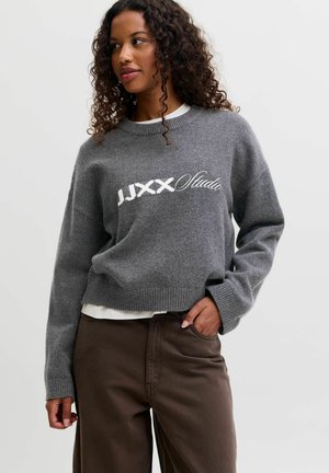 Femme aux cheveux bouclés portant un pull gris avec un texte blanc "JJXX Studio" et un pantalon marron, debout avec une main dans la poche.