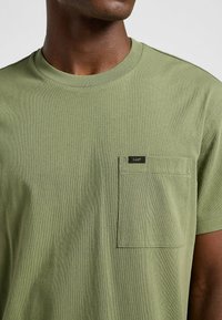 T-shirt in cotone verde oliva con colletto rotondo, maniche corte e taschino sul lato sinistro con un'etichetta nera. Tessuto morbido.
