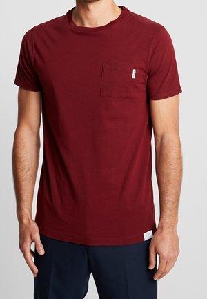 Mężczyzna ubrany w jednolitą bordo koszulkę z krótkim rękawem z małą kieszonką na piersi oraz ciemnoniebieskie spodnie na jasnym tle.