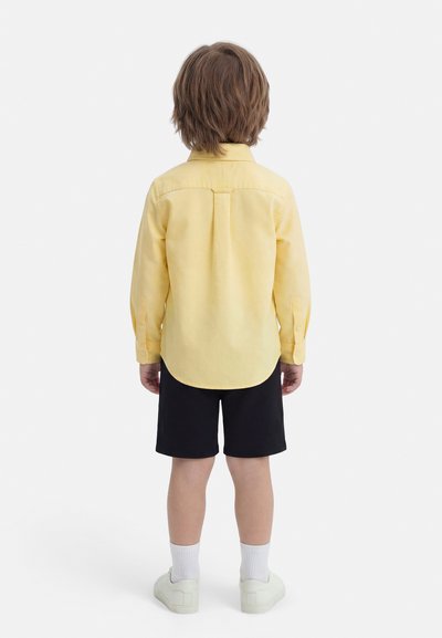 Niño con cabello castaño claro de espaldas, vistiendo una camiseta de manga larga amarilla, pantalones cortos negros, calcetines blancos y zapatillas blancas sobre un fondo blanco.