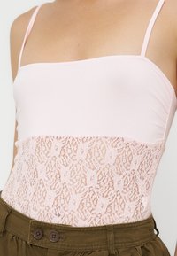 Femme portant un haut sans manches rose pâle avec des détails en dentelle à la taille, associé à un pantalon marron taille haute avec des boutons.