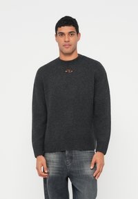 Νεαρός άνδρας που φοράει ένα σκούρο γκρι πλεκτό πουλόβερ με μια μικρή εγκοπή και λογότυπο κοντά στη γραμμή του λαιμού, συνδυασμένο με ξεθωριασμένο μαύρο τζιν.