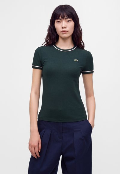 Femme aux cheveux noirs ondulés portant un chemisier vert à manches courtes avec bordure blanche et un pantalon plissé bleu marine, debout avec une main dans la poche.